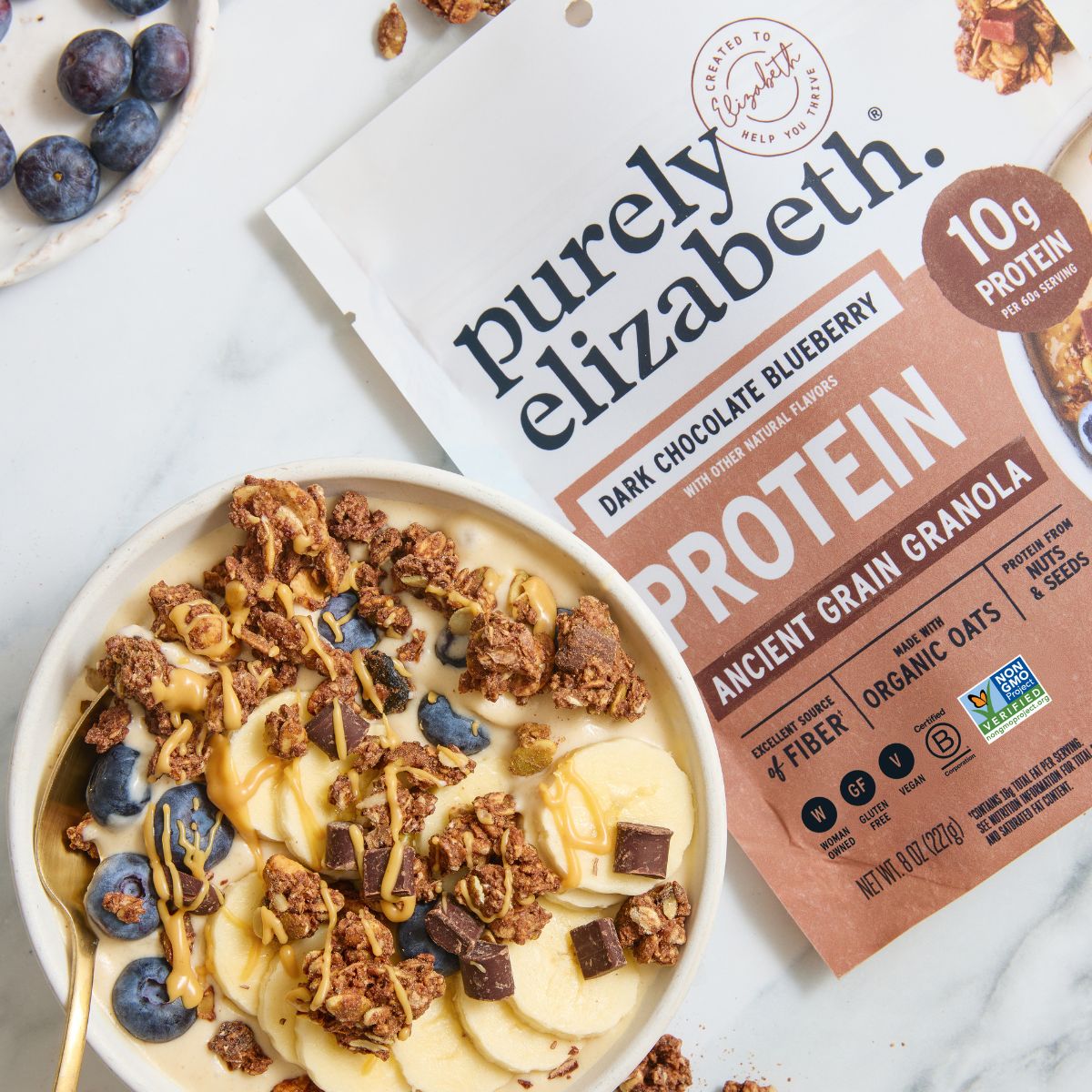 Purely Elizabeth dark chocolate blueberry protein granola bag beside a bowl of granola with blueberries