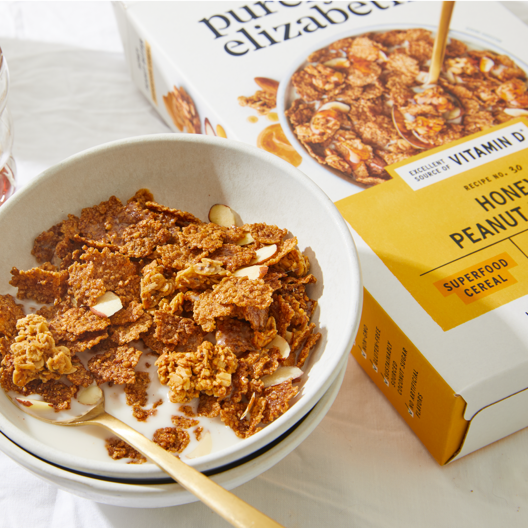 Bowl of crispy granola clusters with milk and a gold spoon, beside a Honey Peanut Butter Superfood cereal box.